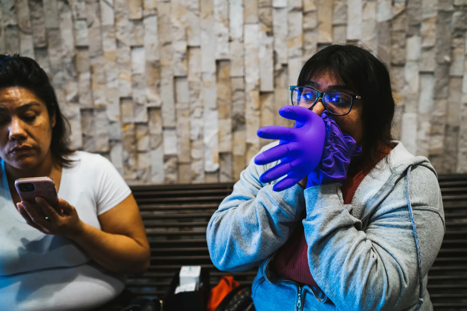 Angie looks to the side as she blows air into a purple medical glove, making it into a balloon. Melissa, out of focus, sits next to her holding a smartphone.