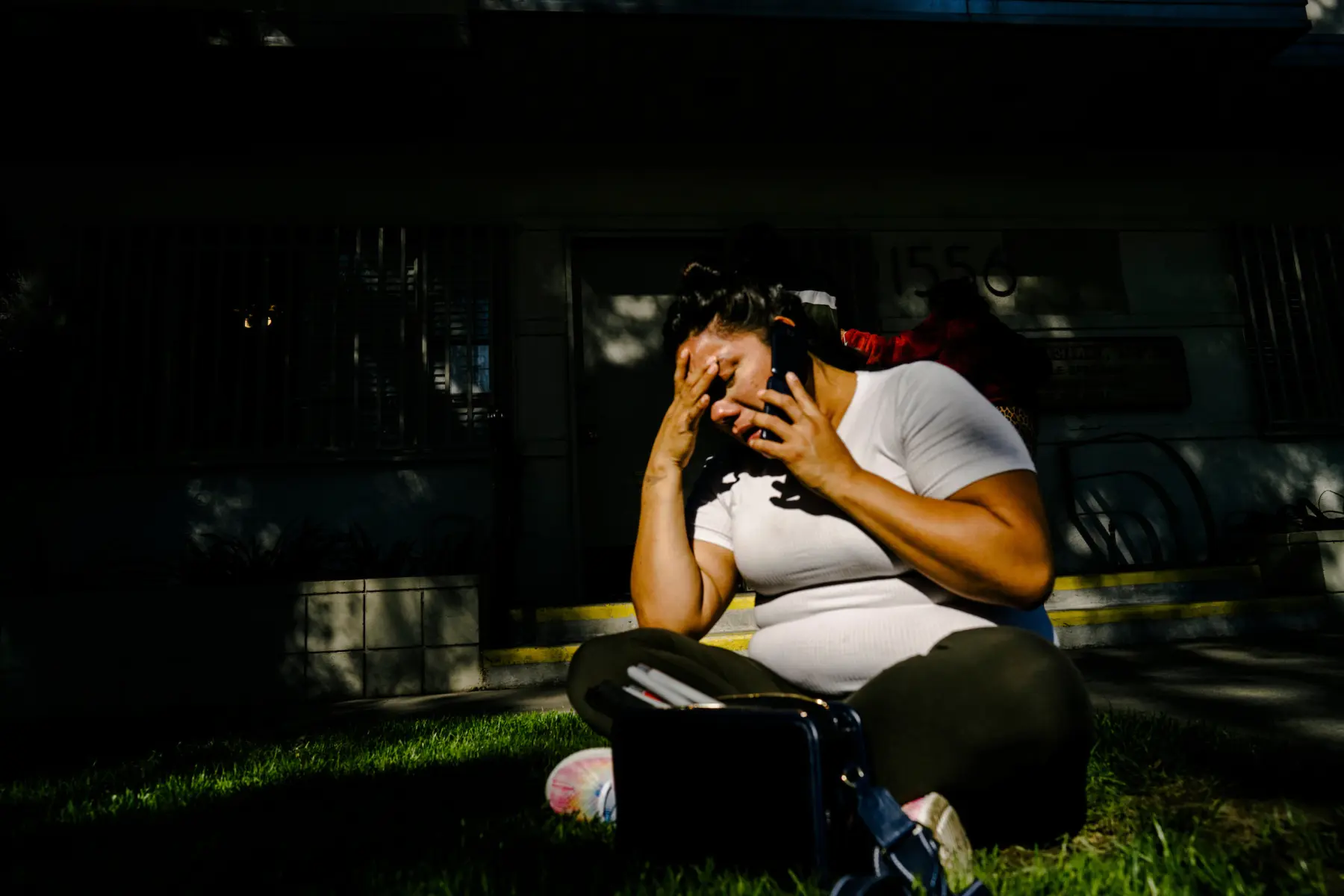 Melissa who sits cross legged on a patch of grass in front of a doctor’s office. She holds her phone to her ear and rests her head on her hand