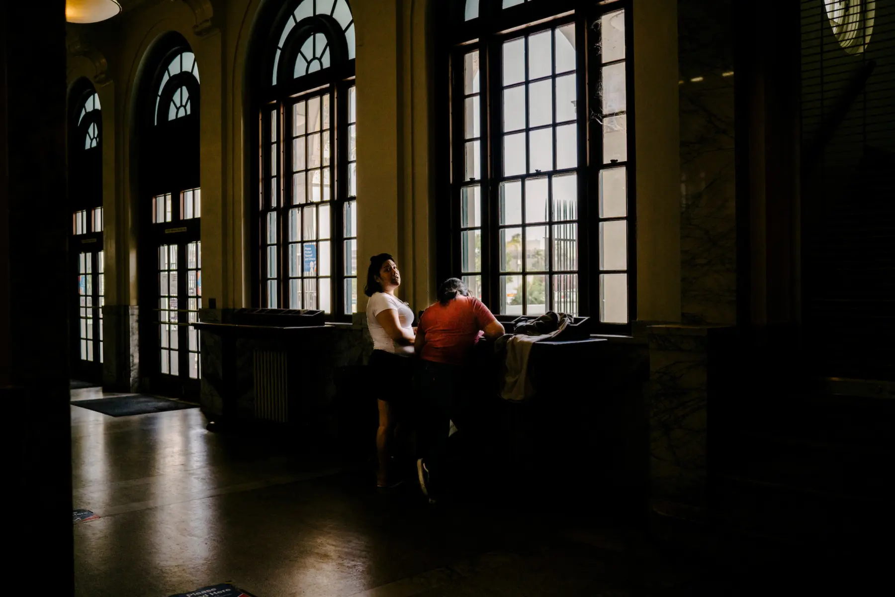 Melissa stands next to Angie in front of a counter at a dimly lit old post office with large glass windows. Melissa is turned toward Angie, who leans against the counter with her head down.