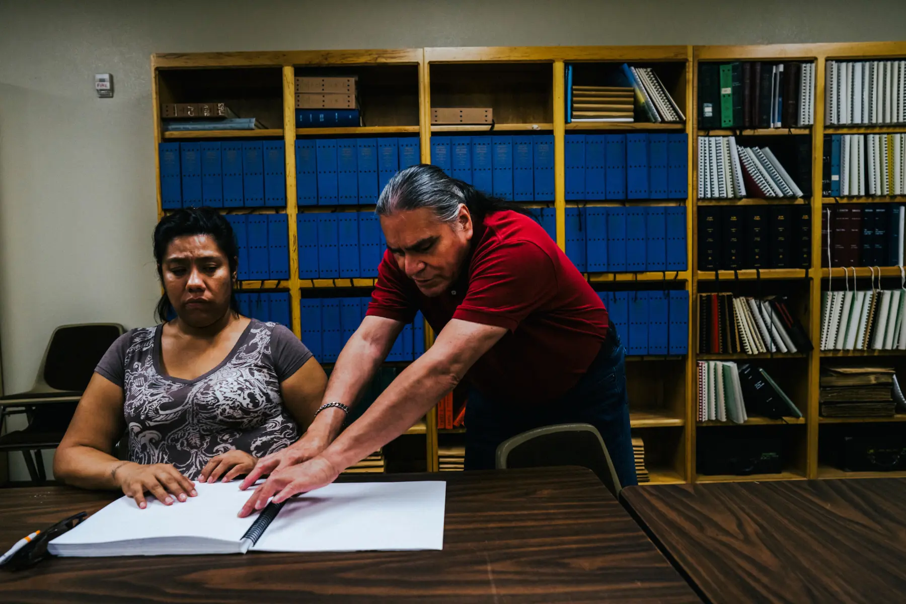 Melissa sits at a conference table with hands on her Braille book. Arturo, an older man with a gray ponytail who is also blind, reads Braille from the same page as he stands next to Melissa. A large case of books is behind them.