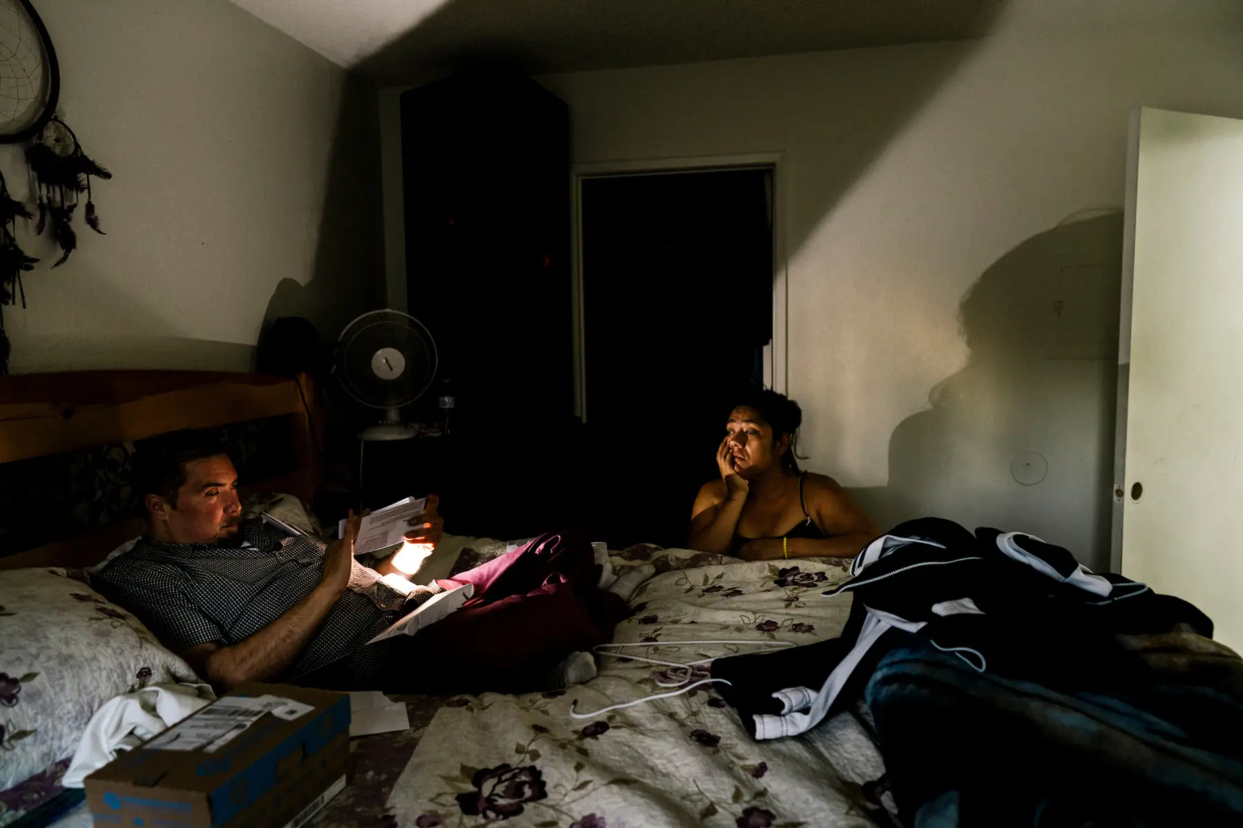Melissa kneels next to a bed while Scott points a smartphone app with flashlight onto a document as he sits on the bed. The flashlight creates a large shadow of Melissa on the white wall behind her.