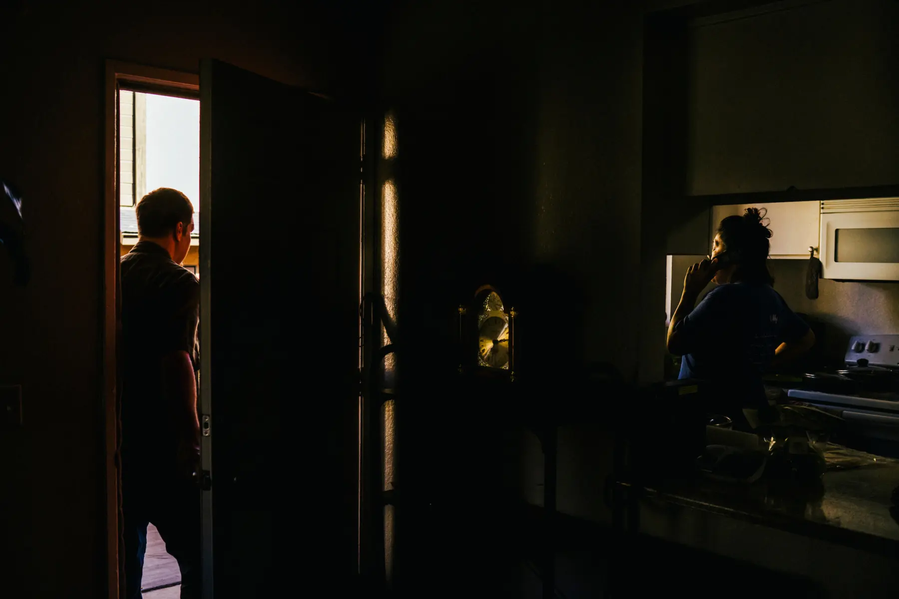 Melissa stands in the dimly lit kitchen on the phone. Scott stands in the front doorway. They are divided by a wall with a large classic clock displayed on a table in front of the wall.