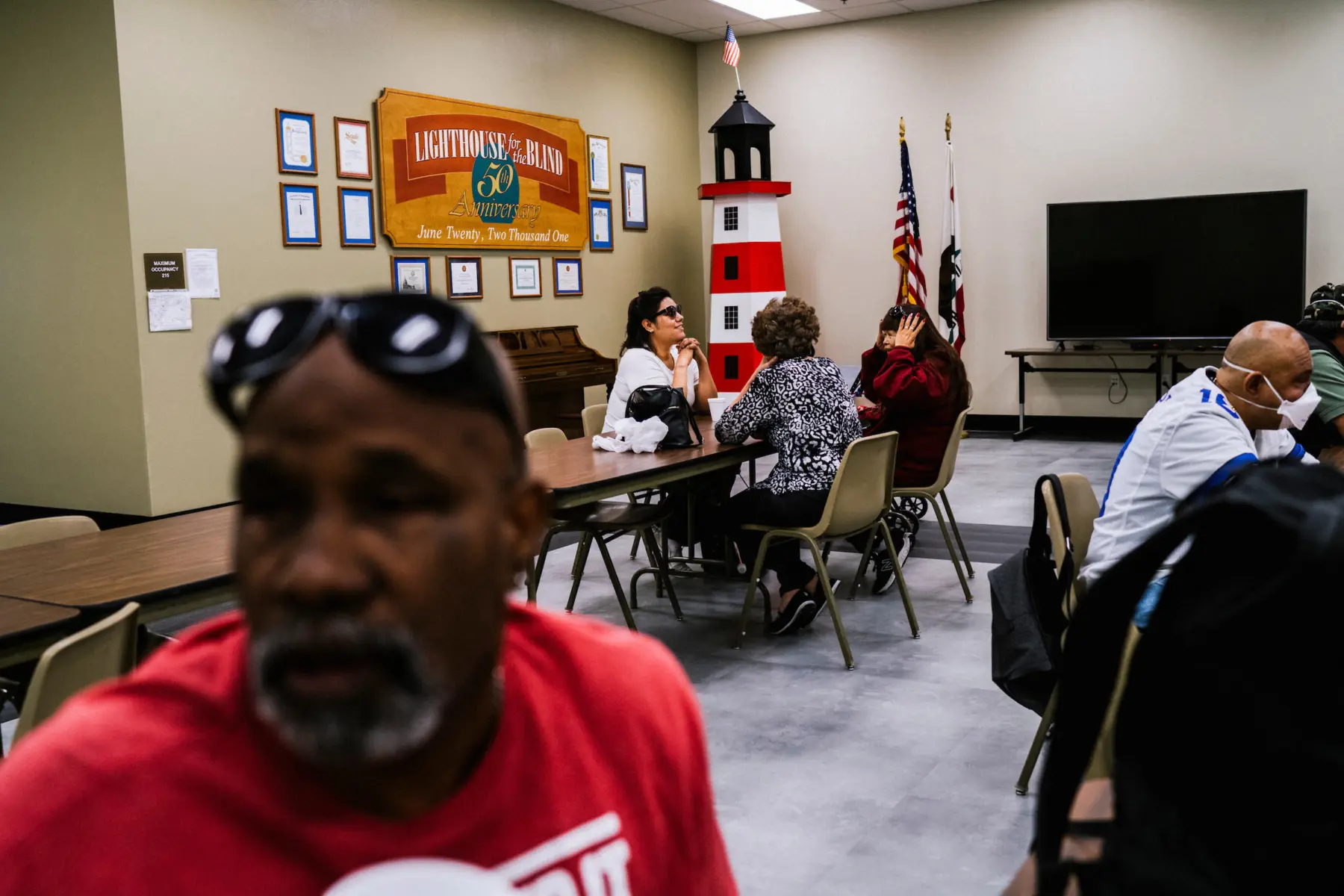 Melissa smiles as she sits with fingers interlaced below her chin at a long table across from two women. In the foreground, a man is out of focus at an adjacent table. Beside him two men sit at the same table. In the faded background, a wooden poster above a piano reads “Lighthouse for the Blind.” A statue of a lighthouse is in the corner next to American and California flags and a television that is turned off.