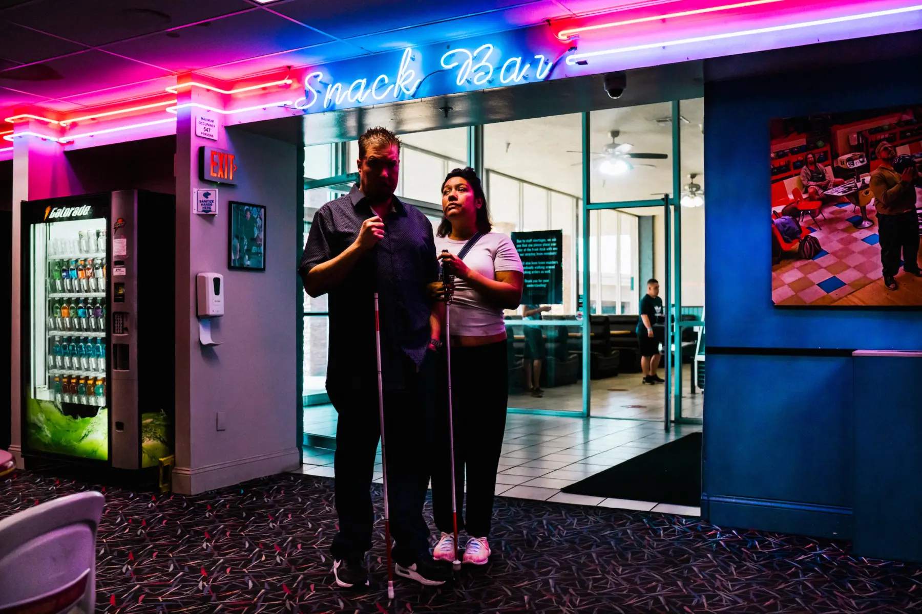 Melissa and Scott stand arm in arm holding their white canes beneath red, pink, and blue neon lights that illuminate their faces. The sign above them reads “snack bar.” A gatorade machine is on the left, and a poster of a man holding a bowling ball is on the right.