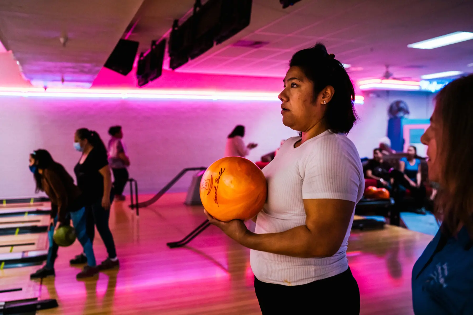 Melissa holds a bowling ball in front of a lane at a bowling alley and a woman who is out of focus is seen on the right looking over at Melissa. People who are out of focus bowl behind Melissa. There are pink neon lights in the background.