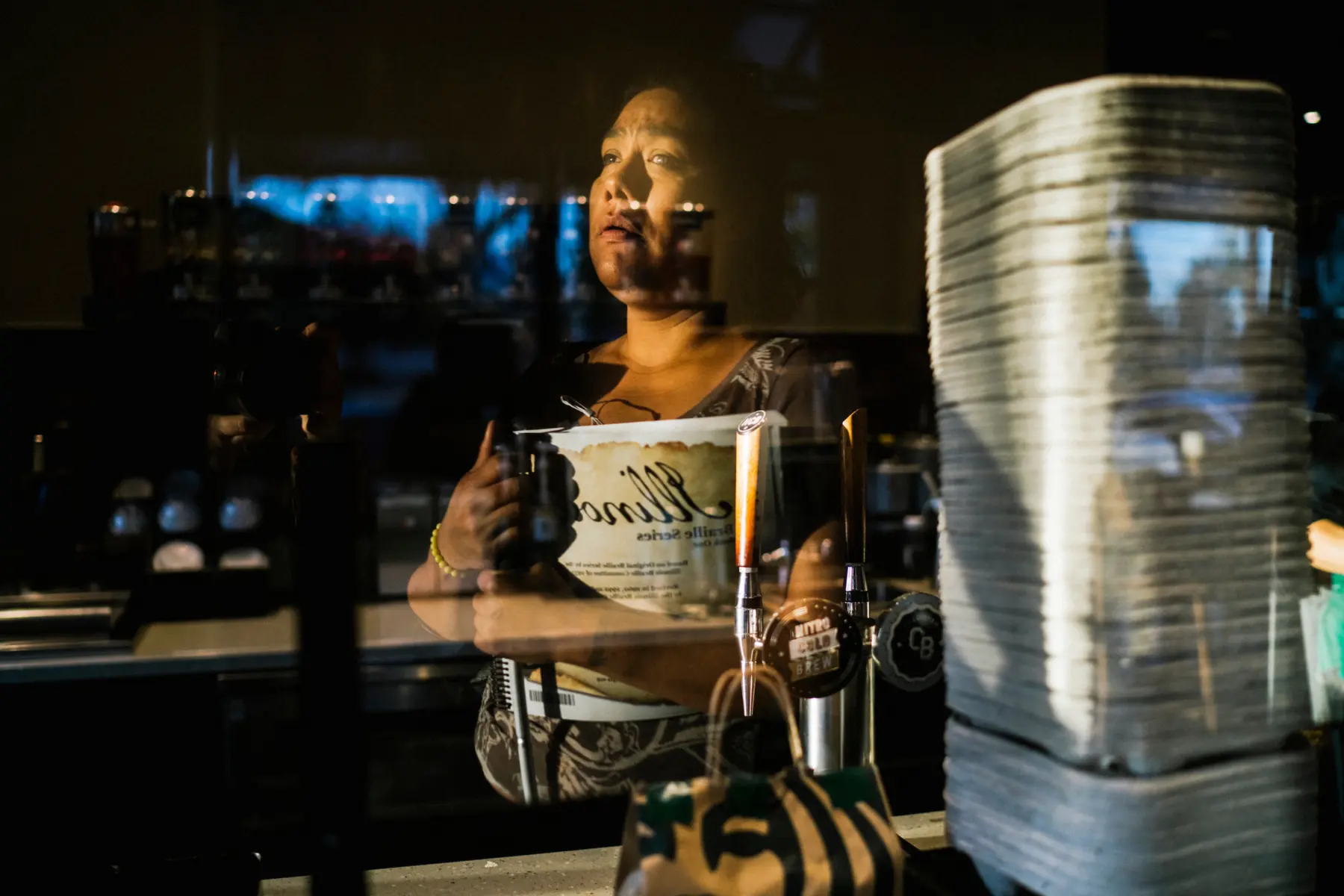 Melissa is reflected in a glass divider at Starbucks coffee. She holds her Braille book to her chest as morning light shines on her face and torso.