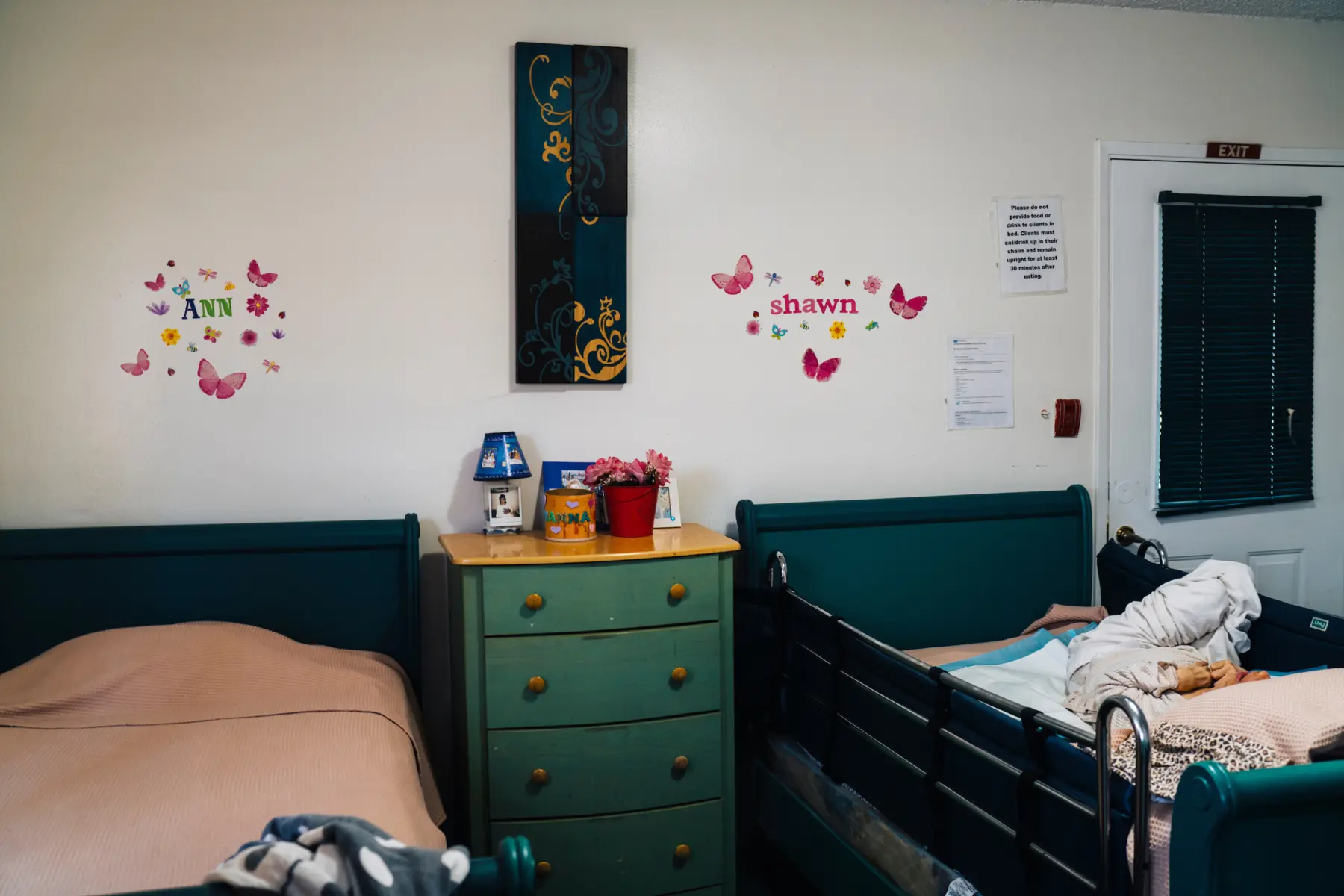 Two beds are on either side of a dresser that holds a small lamp, a Polaroid, a picture frame, and a flower pot. Ann’s bed is empty and her roommate Shawn lies in her own bed. On the wall above Ann’s bed, a sticker collage reads “Ann.” Above her roommate’s bed, stickers read “Shawn.”