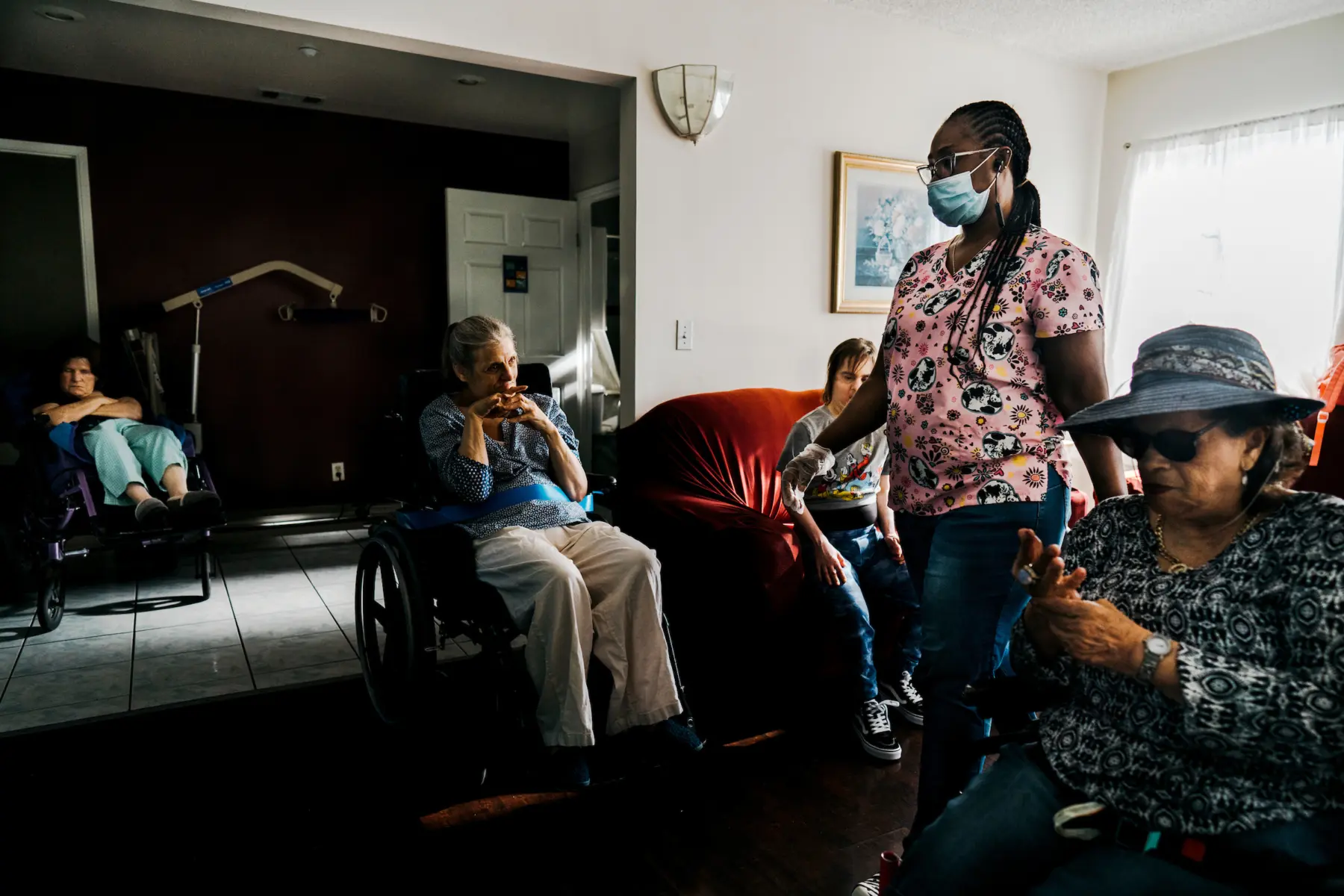 Ann sits in her wheelchair in a living room beside Juliet, a white woman in her 40s, who sits on a red velvet couch. Ann looks to Queen, a Black woman who wears scrubs and a surgical mask, walking by. In front of Queen, Norberta, a Hispanic woman in her 60s in a sunhat and large black sunglasses, sits in her wheelchair massaging her hands. In the far corner  Jill, a white woman in her 50s, reclines in her wheelchair with eyes closed and arms crossed.