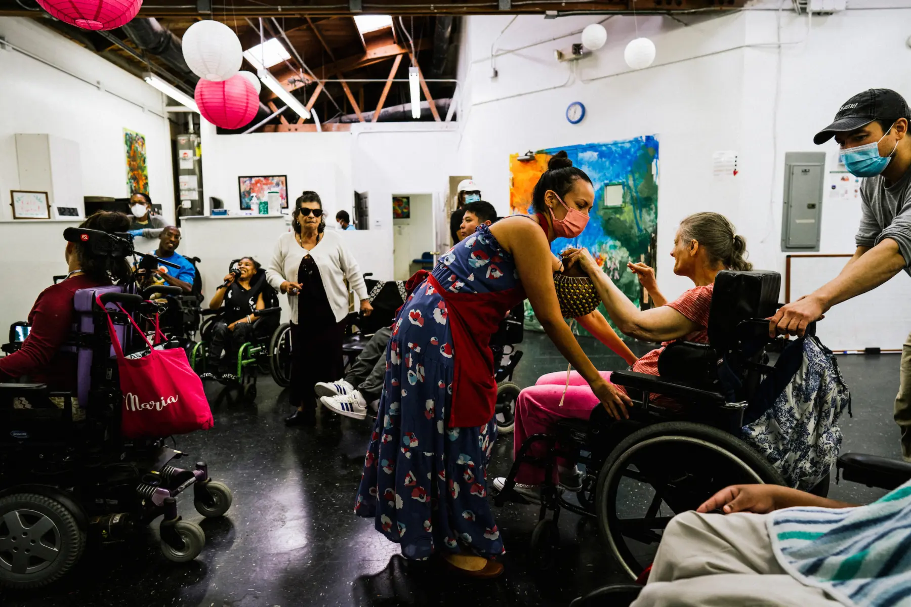 Stephanie, a Hispanic woman in her 30s, pulls Ann forward into group activity at a studio space. Jesse, a biracial Asian male in his 30s, pushes Ann from behind. Ann holds a beaded noise maker as she is maneuvered into a spot among a crowd of other artists who dance, sing, and hold maracas. Art fills the walls and lanterns hang from the ceiling.