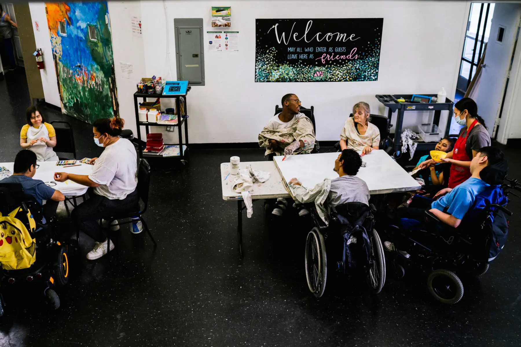 Ann as she sits at a table with a group of artists and an instructor as they paint together on a large white canvas. At another table at left, an instructor sits and draws with two participants. Behind them a poster reads “Welcome, May all who enter as guests leave here as friends.”