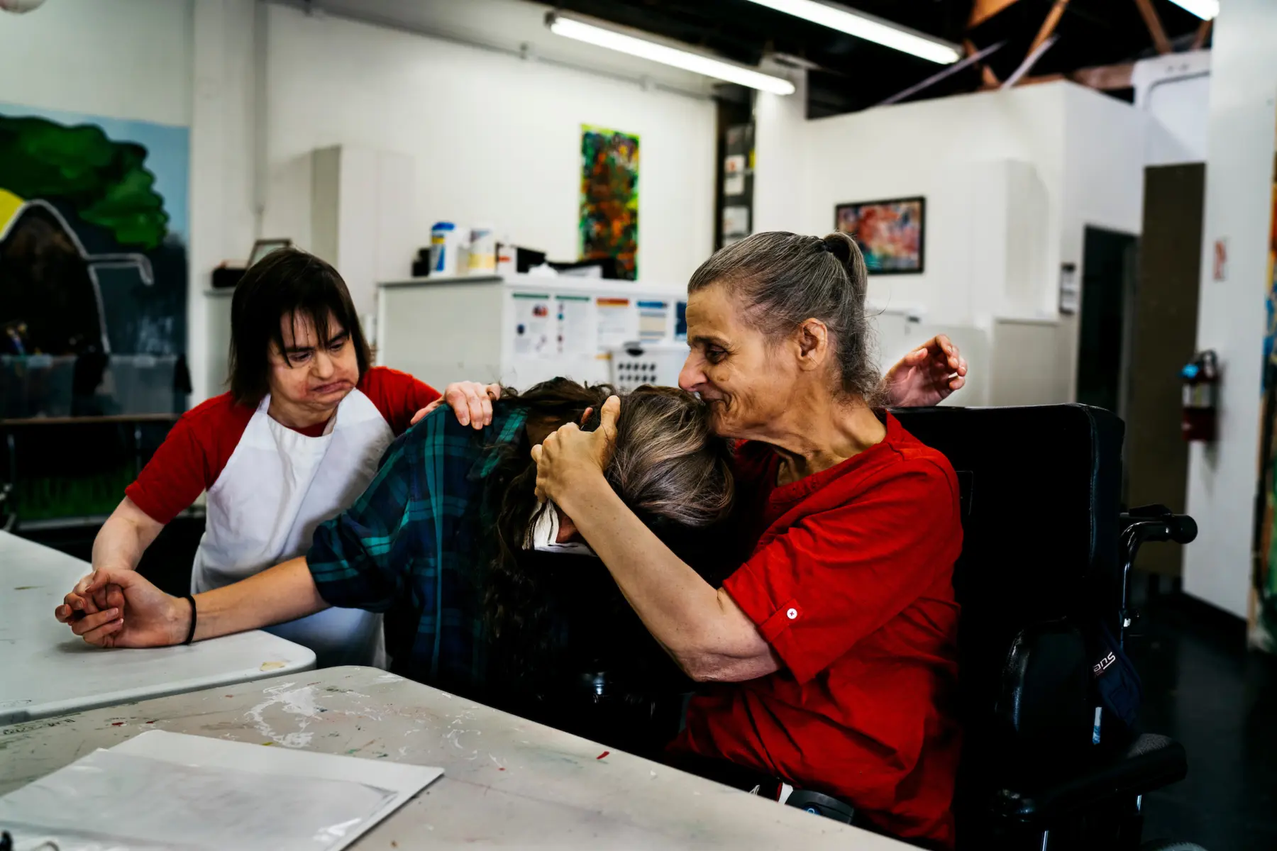 Ann smiles and embraces Dani, a person with brown hair in her 20s, as they sit at a table in an art studio. Juliet, a white woman in her 40s, stands behind Dani with one hand on Dani’s shoulder and another holding Dani’s outstretched hand.