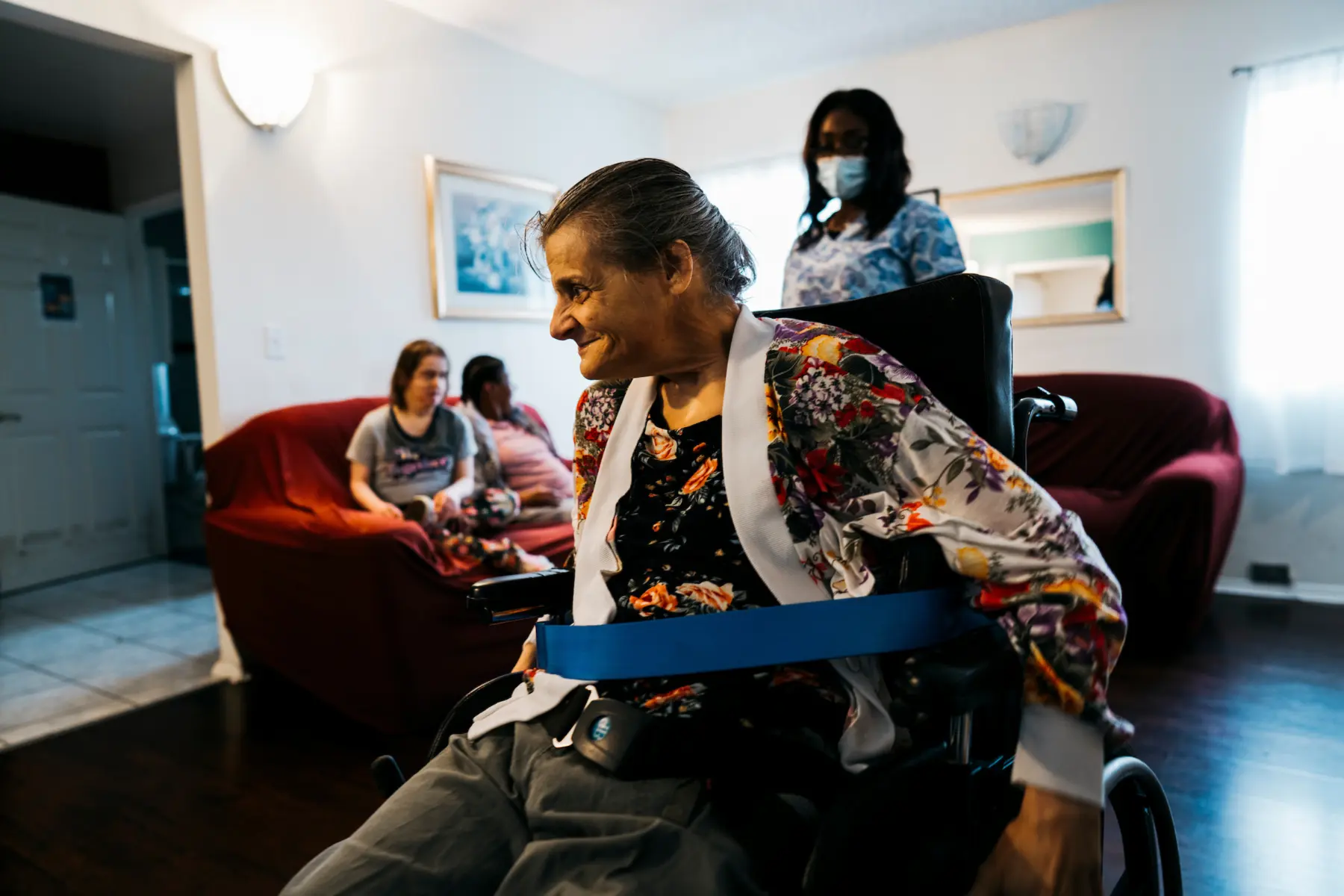 A smiling older woman in a wheelchair, secured with a blue strap, is surrounded by caregivers and others in a cozy living room.