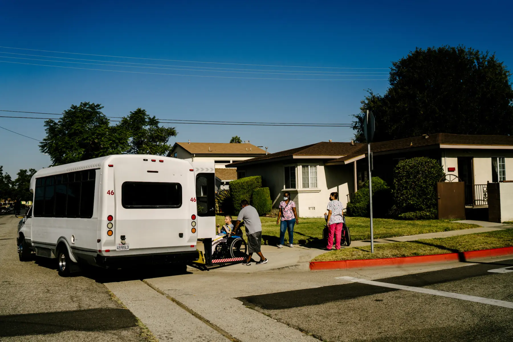 Steve pushes Ann in her wheelchair onto the ramp of a small transport bus parked in front of a single story home on a residential street. Two caregivers in scrubs wait with another resident, who sits in a wheelchair, under a blue sky.