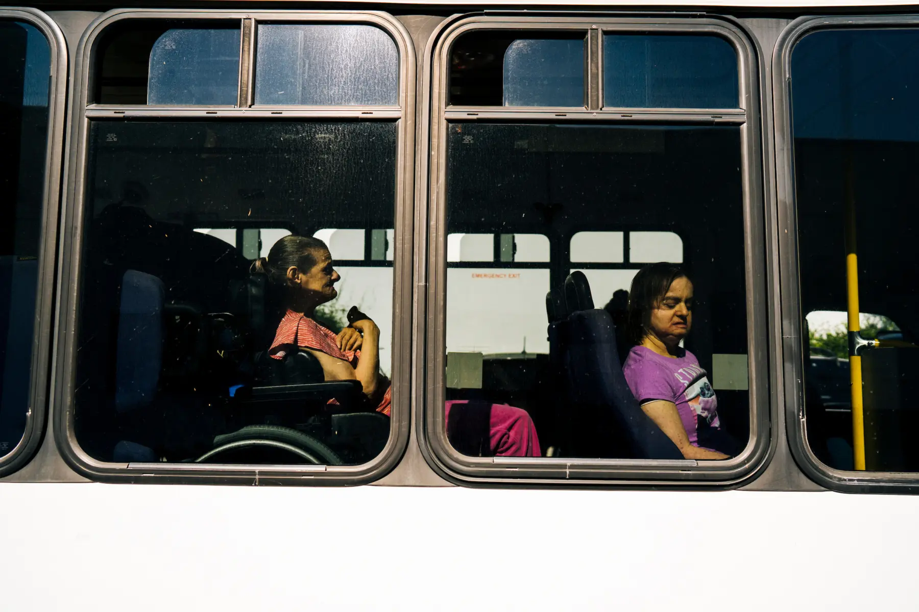 Ann is seen through the tinted windows of a transport bus behind friend Juliet. Sunlight illuminates both.