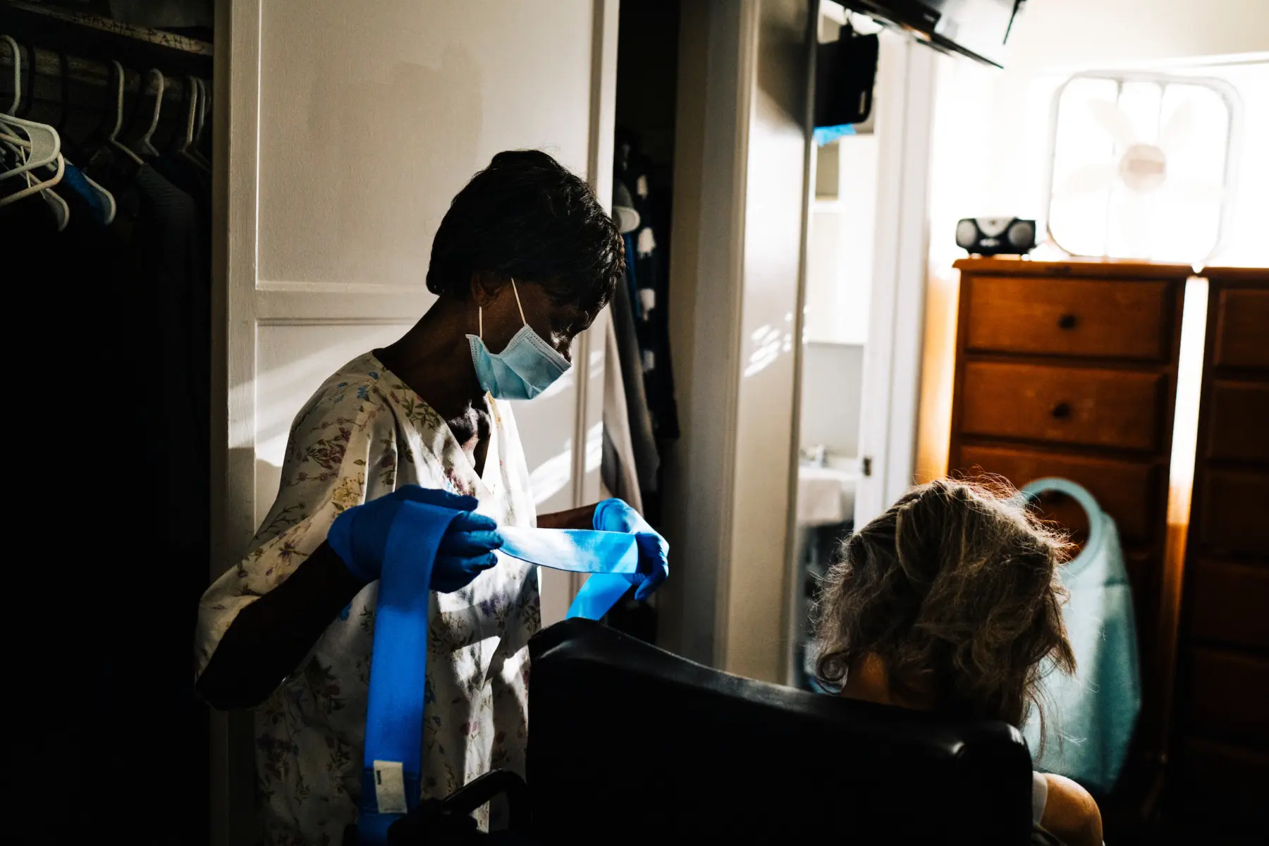Mary, in scrubs and a surgical mask, stands beside Ann in front of Ann’s closet. Ann sits with only the back of her head seen. A soft light shines from the window in front of them, where a dresser with a small boombox and fan on top sits.