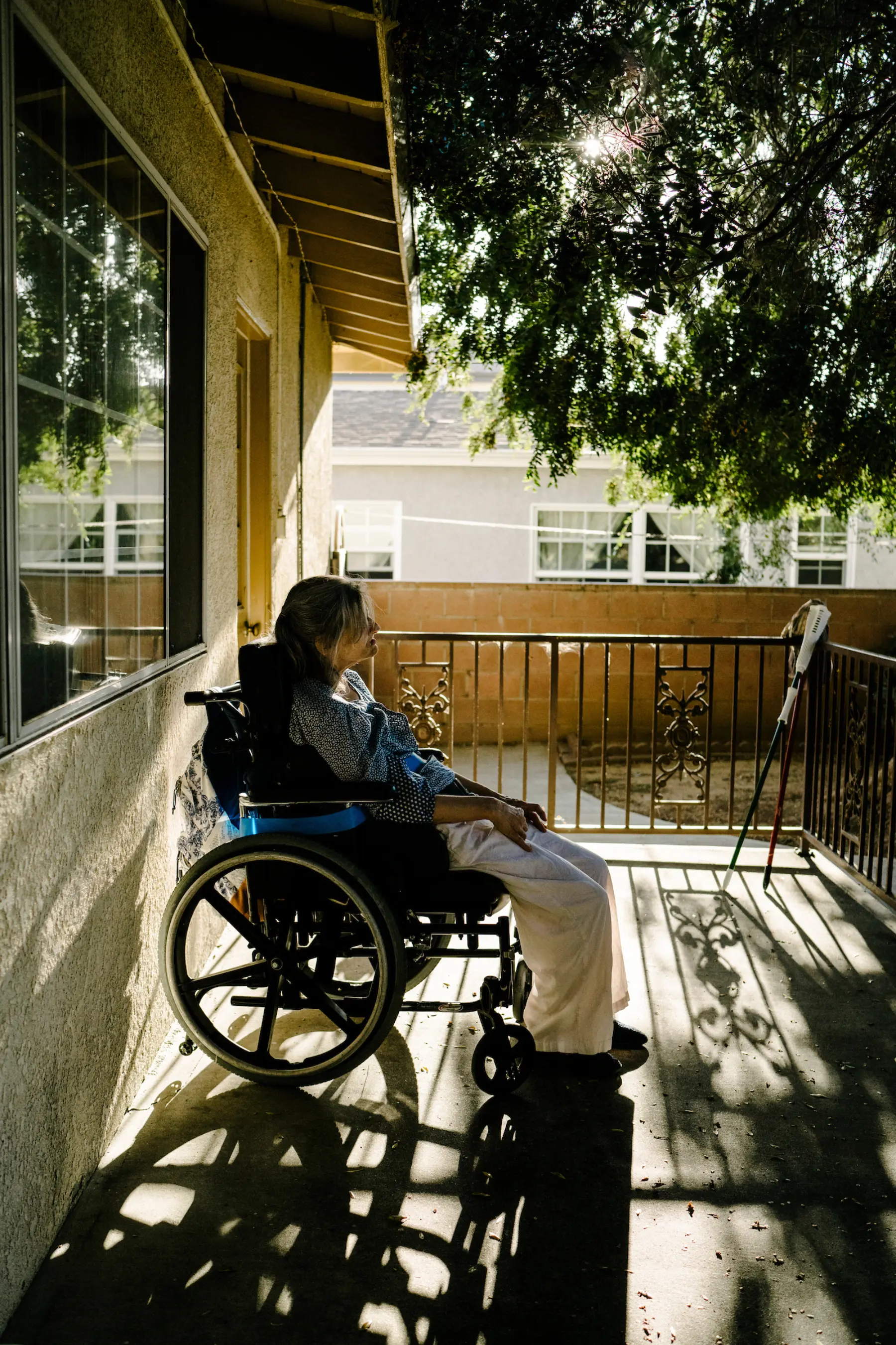 Ann sits on the back patio as light shines through the leaves on a tree. The shadows of a fence and her wheelchair are projected around Ann on the concrete. Another home is seen in the background.