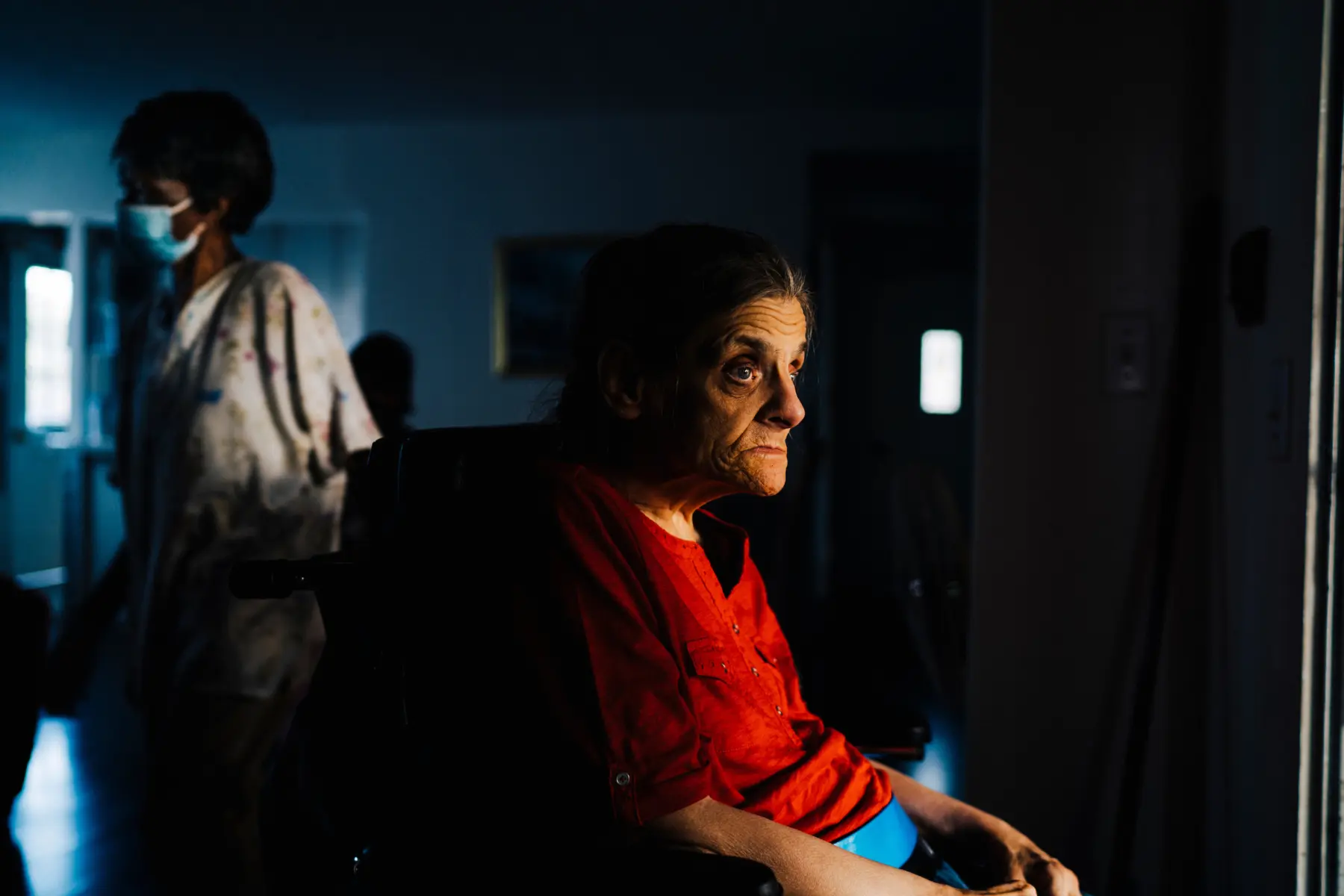A woman in a red shirt sits in a wheelchair, gazing thoughtfully as another figure in the background moves past her.