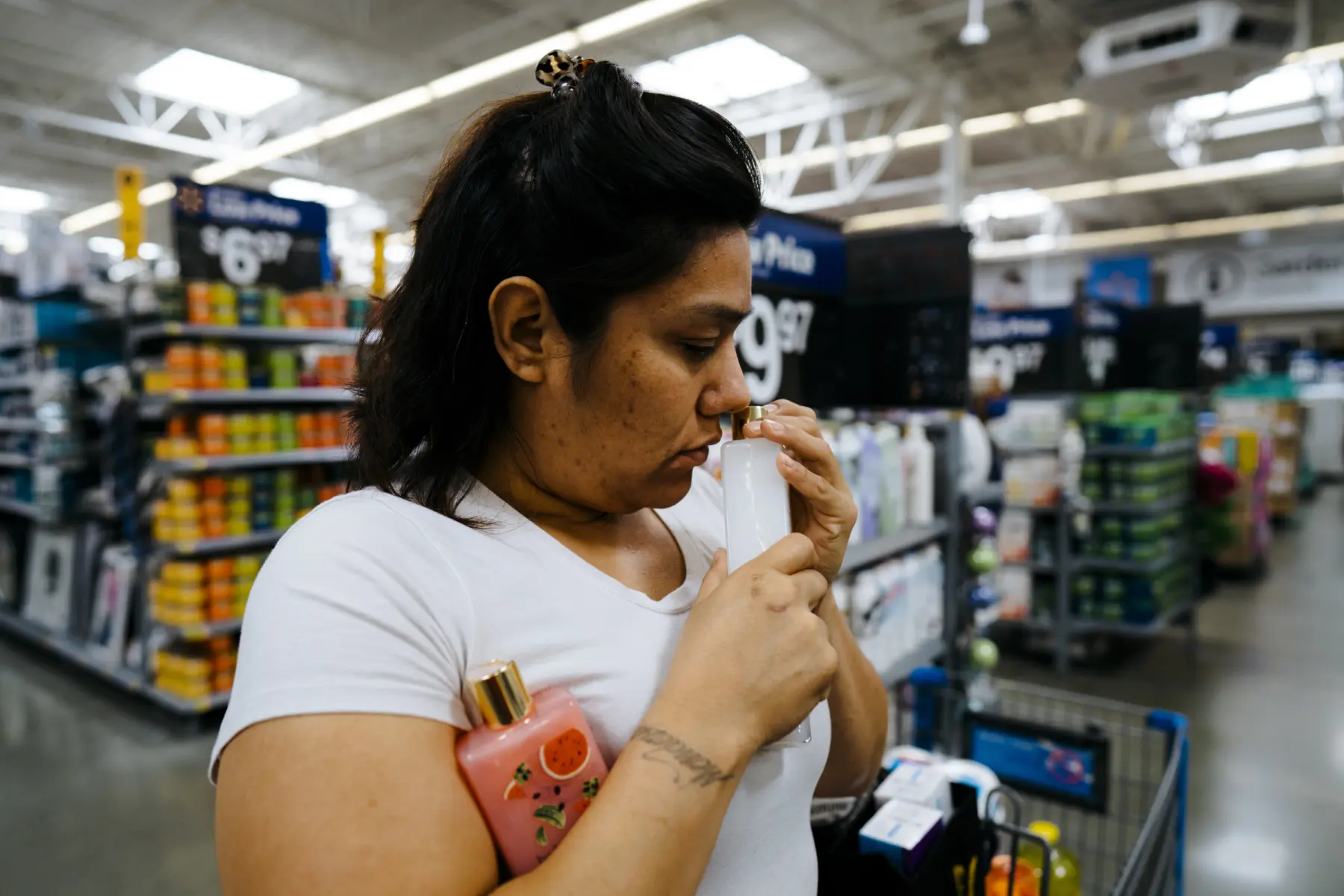 A woman in a white shirt sniffs a bottle of fragrance while holding another brightly colored bottle, surrounded by store shelves.